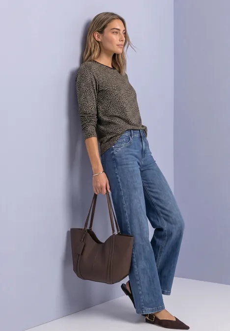 Femme appuyée sur un mur violet, vêtue d’un haut gris, jean large et portant un sac brun.