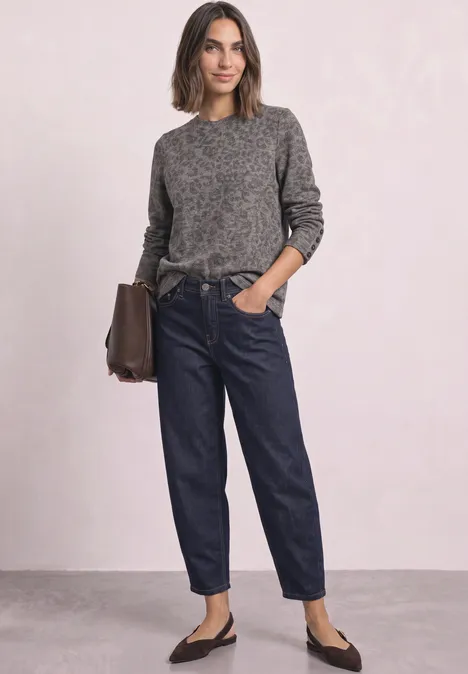 Femme en pull gris, jean bleu foncé, ballerines noires, sac marron, posant devant un mur clair.