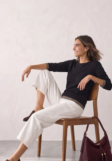 Langarm Shirt mit Colorblock und Printdetail Black Frau in schwarzem Pullover und weißen Hosen sitzt lächelnd auf einem Holzstuhl neben einer Tasche.