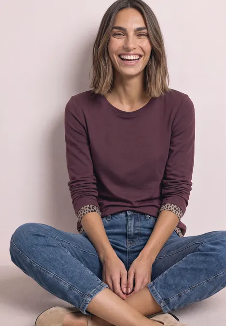 T-shirt à manches longues avec effet color block et imprimé rustic rouge Eine lächelnde Frau in Jeans und weinrotem Langarmshirt sitzt im Schneidersitz vor hellem Hintergrund.