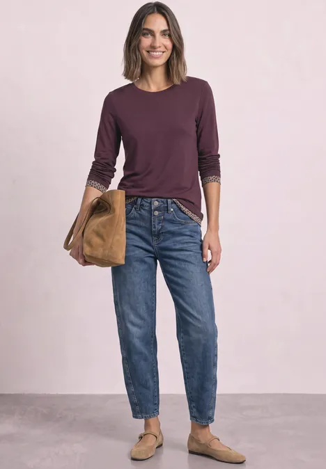 T-shirt à manches longues avec effet color block et imprimé rustic rouge Frau in Jeans, langärmeligem Oberteil, Slippern und Tasche vor hellem Hintergrund.