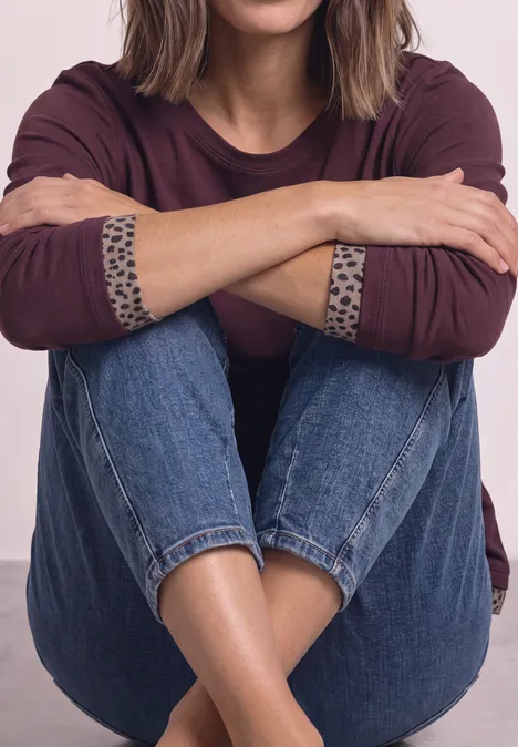 T-shirt à manches longues avec effet color block et imprimé rustic rouge Frau sitzt im Schneidersitz, trägt Jeans und ein dunkelrotes Langarmshirt mit Leo-Bündchen, Arme verschränkt.