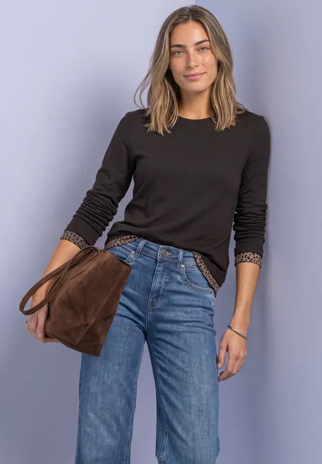 Eine Frau trägt schwarzen Pullover, blaue Jeans und hält eine braune Tasche vor violettem Hintergrund.