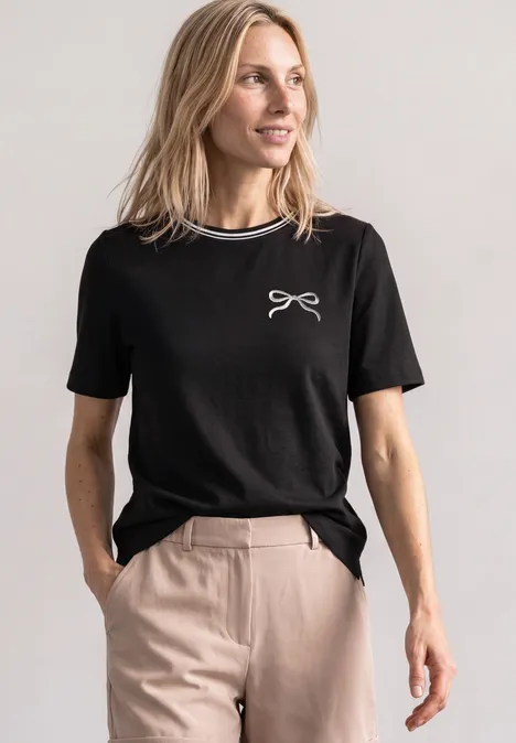 Shirt met ronde hals en geborduurd detail Black Frau in schwarzem T-Shirt mit weißem Schleifen-Print, beiger Hose, Hände in den Taschen, blickt zur Seite.