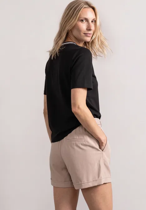 Shirt met ronde hals en geborduurd detail Black Frau mit blondem Haar in schwarzem T-Shirt und beigefarbenen Shorts steht seitlich, eine Hand in der Tasche.