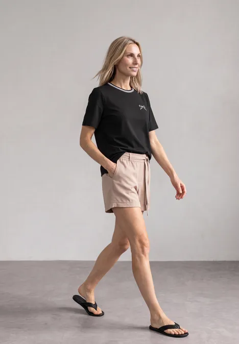 Shirt met ronde hals en geborduurd detail Black Frau in schwarzem Shirt, beigen Shorts und Flipflops schreitet gelassen.