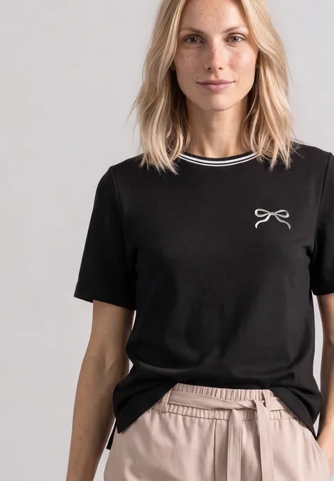 Shirt met ronde hals en geborduurd detail Black Eine Frau trägt ein schwarzes T-Shirt mit kleinem Schleifendruck und beigen Hosen vor neutralem Hintergrund.