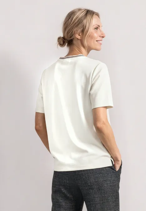 Frau in weißem Kurzarm-Pullover und grauer Hose, seitlich stehend und lächelnd vor hellem Hintergrund.