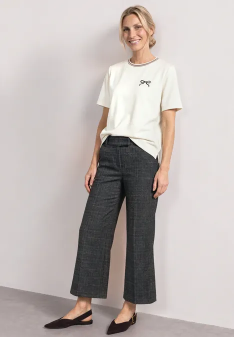 Frau trägt weißes T-Shirt, weite Karohose und schwarze Ballerinas vor hellem Hintergrund.