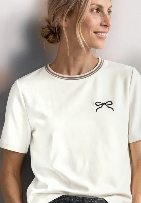 Eine lächelnde Frau mit blondem Dutt trägt ein weißes T-Shirt mit dünnem Ringel-Kragen und kleinem Schleifen-Print.