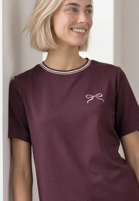 Shirt met ronde hals en geborduurd detail rustic rouge Een jonge vrouw in een bordeaux T-shirt met witte boord en klein strikmotief, glimlachend.