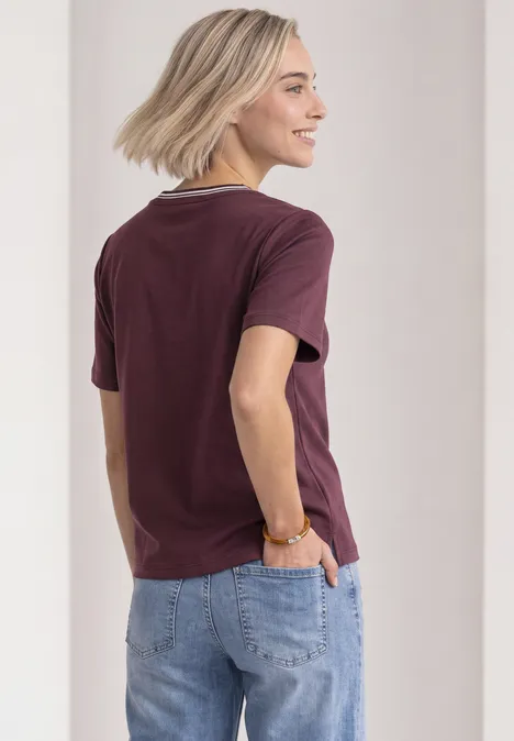 Shirt met ronde hals en geborduurd detail rustic rouge Een vrouw met kort blond haar draagt een bordeauxrood shirt en een blauwe spijkerbroek, gezien van achteren.