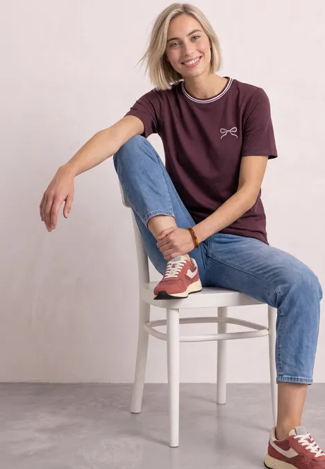 Shirt met ronde hals en geborduurd detail rustic rouge Een jonge vrouw zit glimlachend op een witte stoel, met jeans, bordeaux T-shirt en sneakers.