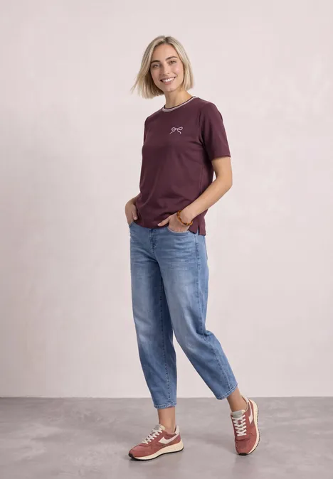 Shirt met ronde hals en geborduurd detail rustic rouge Blonde vrouw in een bordeaux t-shirt, blauwe spijkerbroek en sneakers, glimlachend voor een lichte achtergrond.