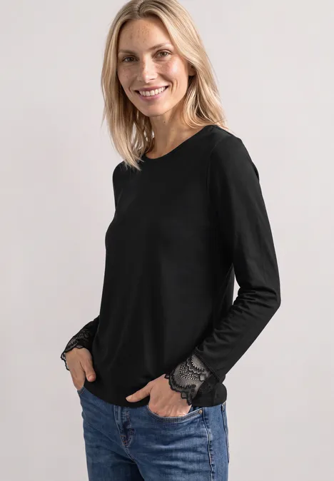 Langarm Shirt mit Rundhals und Spitze Black Eine lächelnde Frau mit hellem Haar trägt einen schwarzen Langarmshirt mit Spitzenbündchen und Jeans.