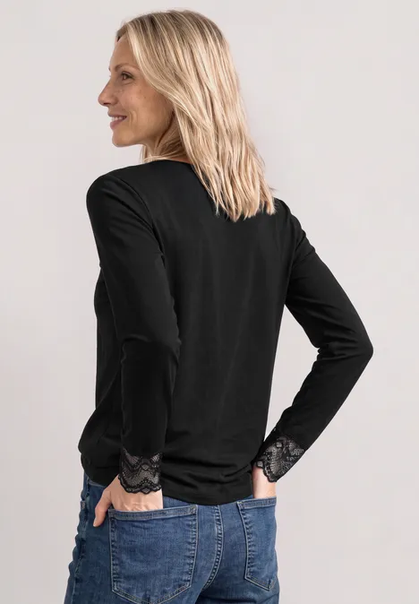 Langarm Shirt mit Rundhals und Spitze Black Seitlich blickende Frau in schwarzem Spitzenshirt und blauen Jeans vor neutralem Hintergrund.