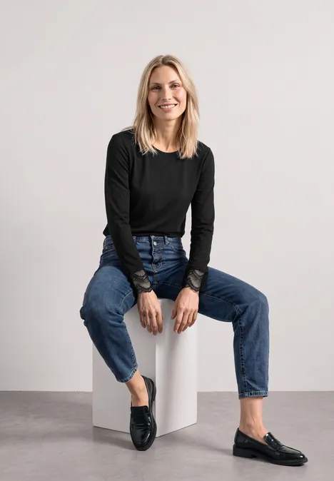 Langarm Shirt mit Rundhals und Spitze Black Eine blonde Frau sitzt auf einem weißen Würfel, trägt eine schwarze Bluse, Jeans und lächelt.