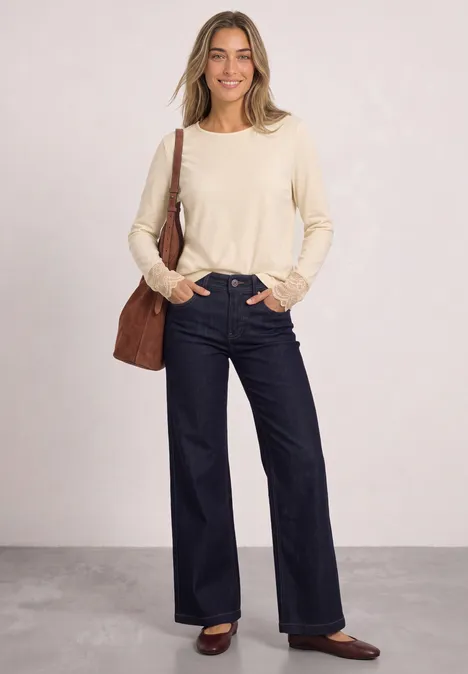 Eine Frau in einem beigen Pullover, weiten dunkelblauen Jeans und braunen Schuhen mit Schultertasche.