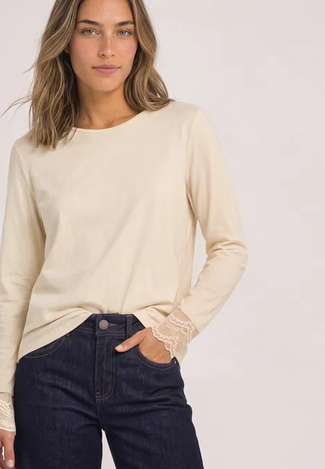Junge Frau vor neutralem Hintergrund trägt hellbeiges Langarmshirt und blaue Jeans, linke Hand in Tasche.