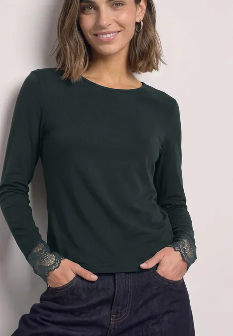 Langarm Shirt mit Rundhals und Spitze stormy green Frau trägt dunkelgrünes Langarmshirt mit Spitzenmanschetten und blauen Jeans, lächelnd mit Händen in den Taschen.