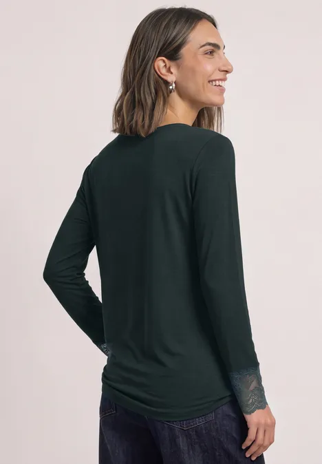 Langarm Shirt mit Rundhals und Spitze stormy green Frau in dunkelgrüner Langarmbluse mit Spitzendetails an den Ärmeln, von hinten seitlich abgelichtet.