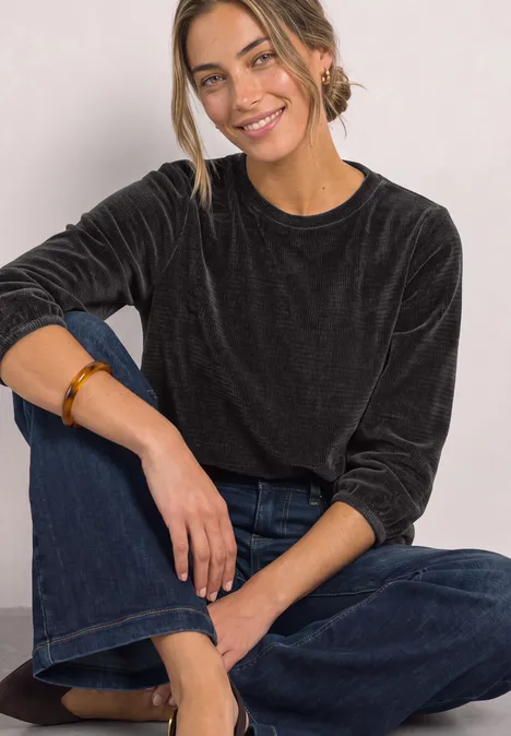 Junge Frau sitzt entspannt auf dem Boden, trägt schwarzen Samt-Pullover und weite Jeans, lächelt.