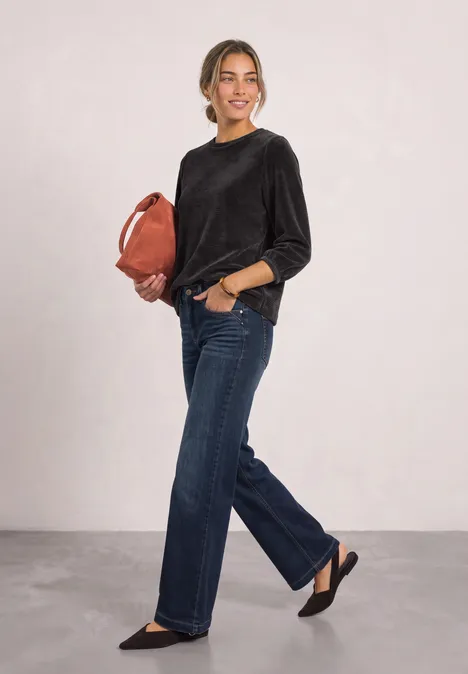 Frau im schwarzen Pullover und weiten Jeans, trägt eine braune Tasche und schwarze Ballerinas vor hellem Hintergrund.