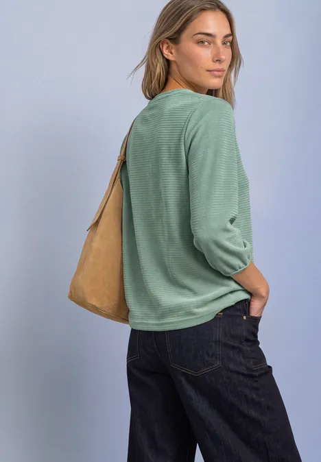 Eine Frau in lässigem grünem Pullover, Jeans und brauner Schultertasche blickt über die Schulter.