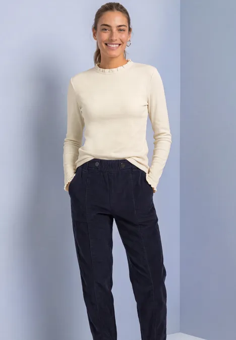 Langarm Shirt mit Rundhals und Rüschen pure beige Frau lächelt, trägt cremefarbenes Langarmshirt und schwarze Hose, steht vor hellblauem Hintergrund.