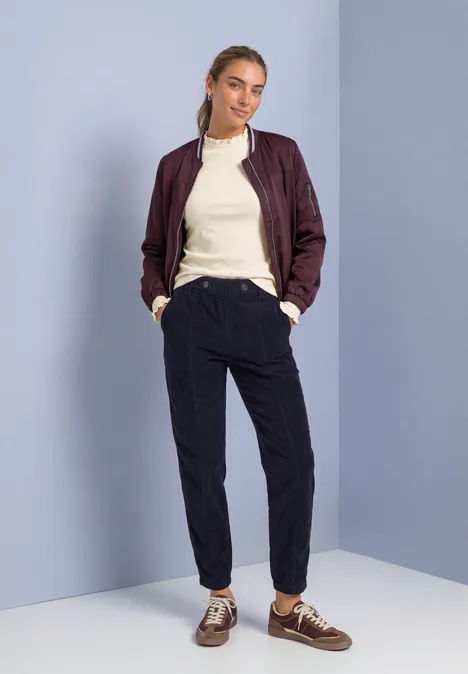 Langarm Shirt mit Rundhals und Rüschen pure beige Model trägt rote Bomberjacke, cremefarbenes Top, schwarze Jogginghose und braune Schuhe, steht in Raumecke.