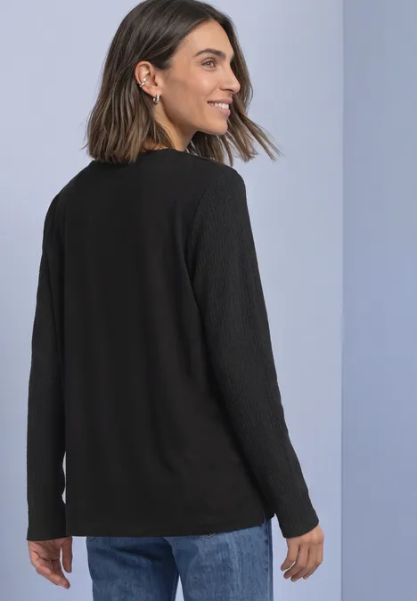 Langarm Shirt mit Struktur und Knöpfen Black Frau blickt lachend über ihre Schulter, trägt einen schwarzen Pullover und Jeans vor hellem Hintergrund.