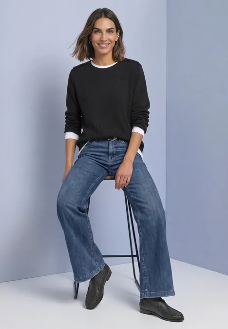 Langarm Shirt mit Struktur und Knöpfen Black Frau sitzt auf Hocker in schwarzem Pullover mit weißem Kragen, weiten blauen Jeans und schwarzen Stiefeln.