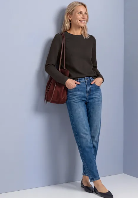 Frau lehnt entspannt an Wand, trägt schwarzen Pullover, blaue Jeans und rote Tasche, lächelt leicht.
