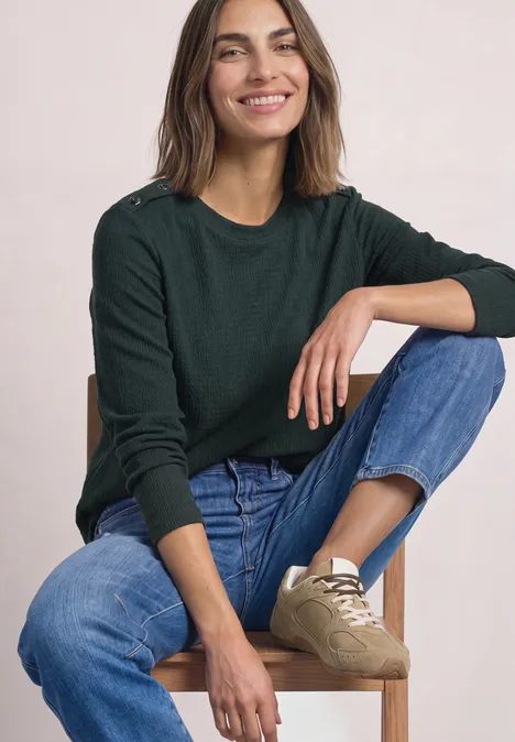 Shirt met lange mouwen, structuur en knopen stormy green Een glimlachende vrouw in een groene trui en spijkerbroek zit ontspannen op een houten stoel.