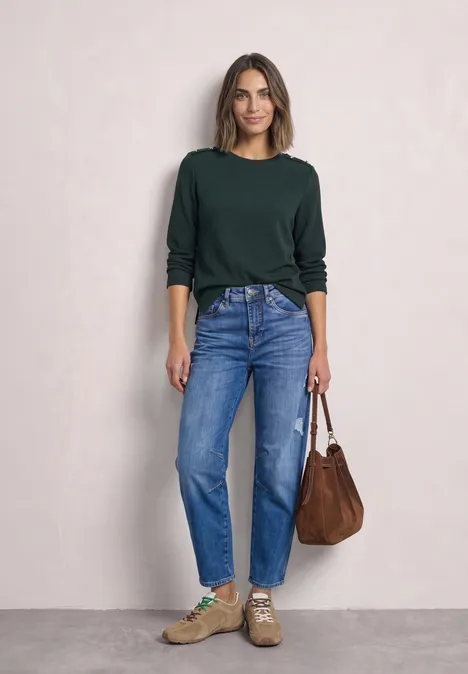 Shirt met lange mouwen, structuur en knopen stormy green Lachende vrouw voor lichte muur in groene trui, blauwe jeans, sneakers en een bruine schoudertas.