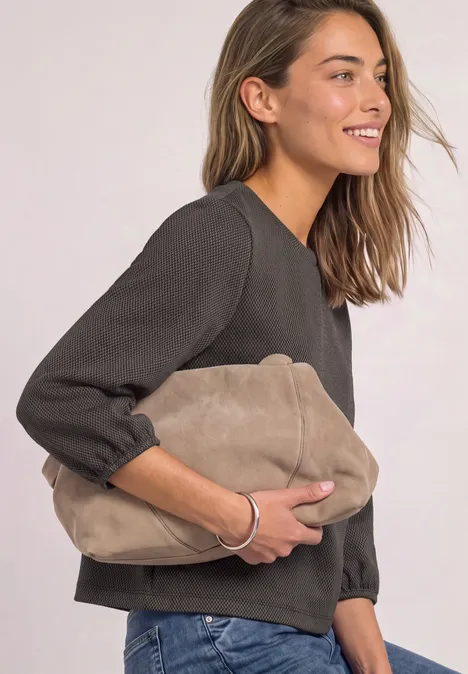 Eine lächelnde Frau hält eine beige Clutchtasche vor hellem Hintergrund.