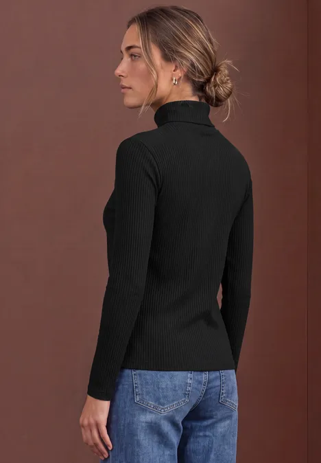 Basic coltrui Black Eine Frau in schwarzem Rollkragenpullover und Jeans steht seitlich vor braunem Hintergrund.