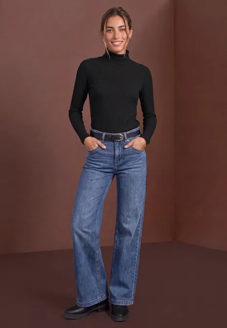 Basic coltrui Black Eine Frau steht vor braunem Hintergrund in schwarzem Rollkragenpullover und ausgestellten Jeans, lächelnd.