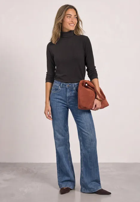 Frau in schwarzem Rollkragenpullover, weiten Jeans und schwarzen Schuhen trägt eine braune Handtasche vor hellem Hintergrund.