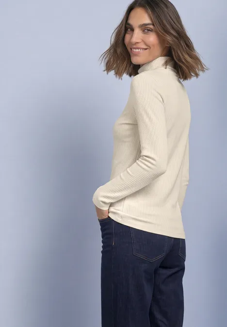 T-shirt basique uni à col roulé pure beige Frau in cremefarbenem Rollkragenpullover und blauen Jeans schaut über die Schulter und lächelt.