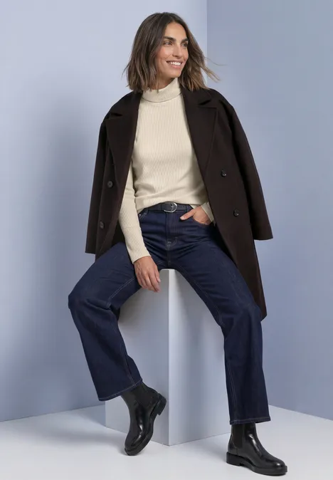 T-shirt basique uni à col roulé pure beige Frau in braunem Mantel, Rollkragenpullover, weiter Jeans und schwarzen Stiefeln sitzt lächelnd auf weißem Block