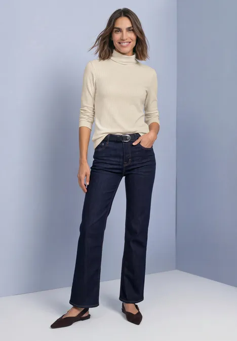 T-shirt basique uni à col roulé pure beige Frau in hellem Rollkragenpullover, dunklen Jeans und schwarzen flachen Schuhen vor schlichtem Hintergrund