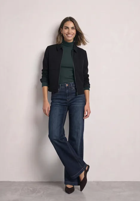 Basic shirt met col stormy green Een vrouw in een zwart jasje, groen shirt en wijde spijkerbroek staat lachend voor een lichte achtergrond.