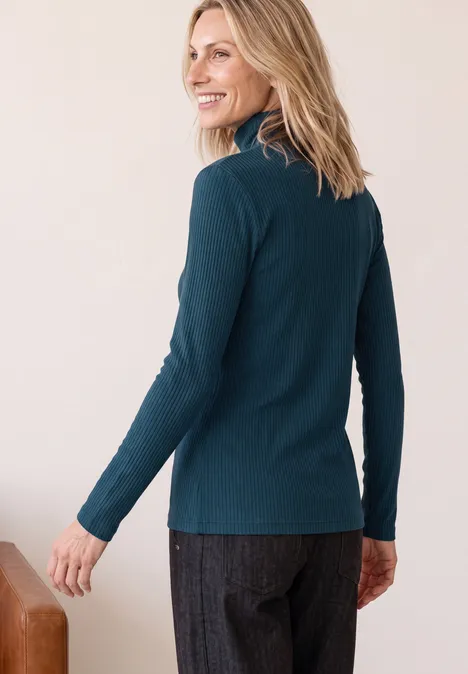 Frau in dunkelgrünem Rollkragenpullover und grauen Hosen, Blick über die Schulter, lächelnd.