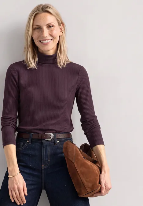 T-shirt basique uni à col roulé berry plum Frau mit blondem Haar, dunkelviolettem Rollkragenpullover und Jeans, hält eine braune Tasche und lächelt.