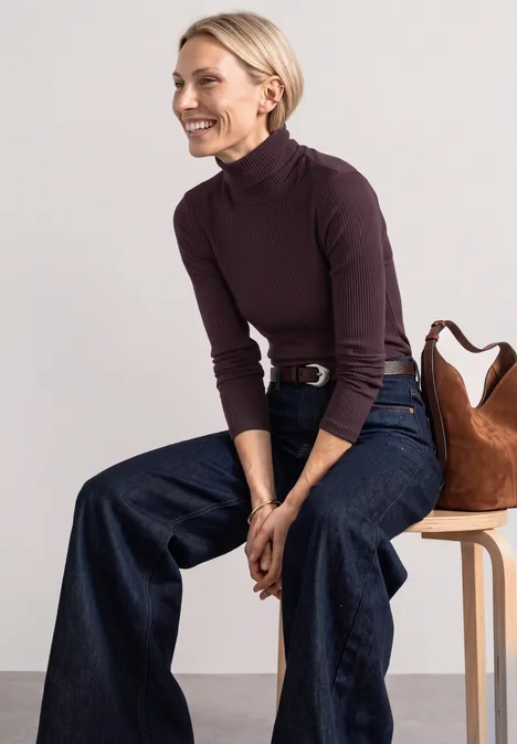 T-shirt basique uni à col roulé berry plum Frau in lila Rollkragenpullover und weiten Jeans sitzt lächelnd auf einem Hocker mit Tasche.