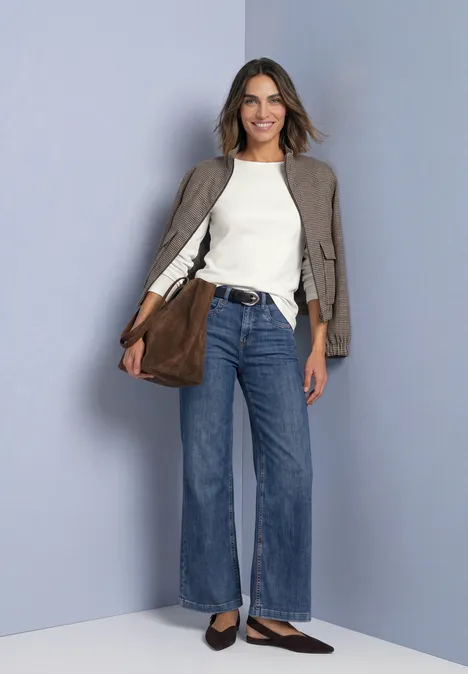 Lachende vrouw in wijde blauwe jeans, wit shirt, taupe jasje en bruine tas voor een grijze achtergrond.