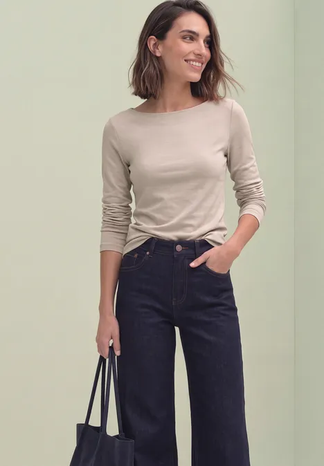 Eine lächelnde Frau in beigefarbenem Langarmshirt, dunklen Jeans mit weitem Bein und schwarzer Tasche