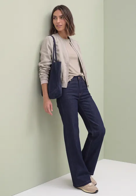 Frau in beigem Pullover, Blazer, weiter Jeans und Tasche lehnt an hellgrüner Wand.