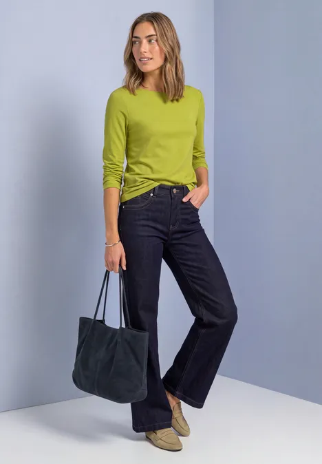 Basic Shirt met boothals apple lime Vrouw in citroengroen shirt met lange mouwen, donkere jeans en beige laarsjes, marineblauwe tas, lichtblauwe achtergrond.